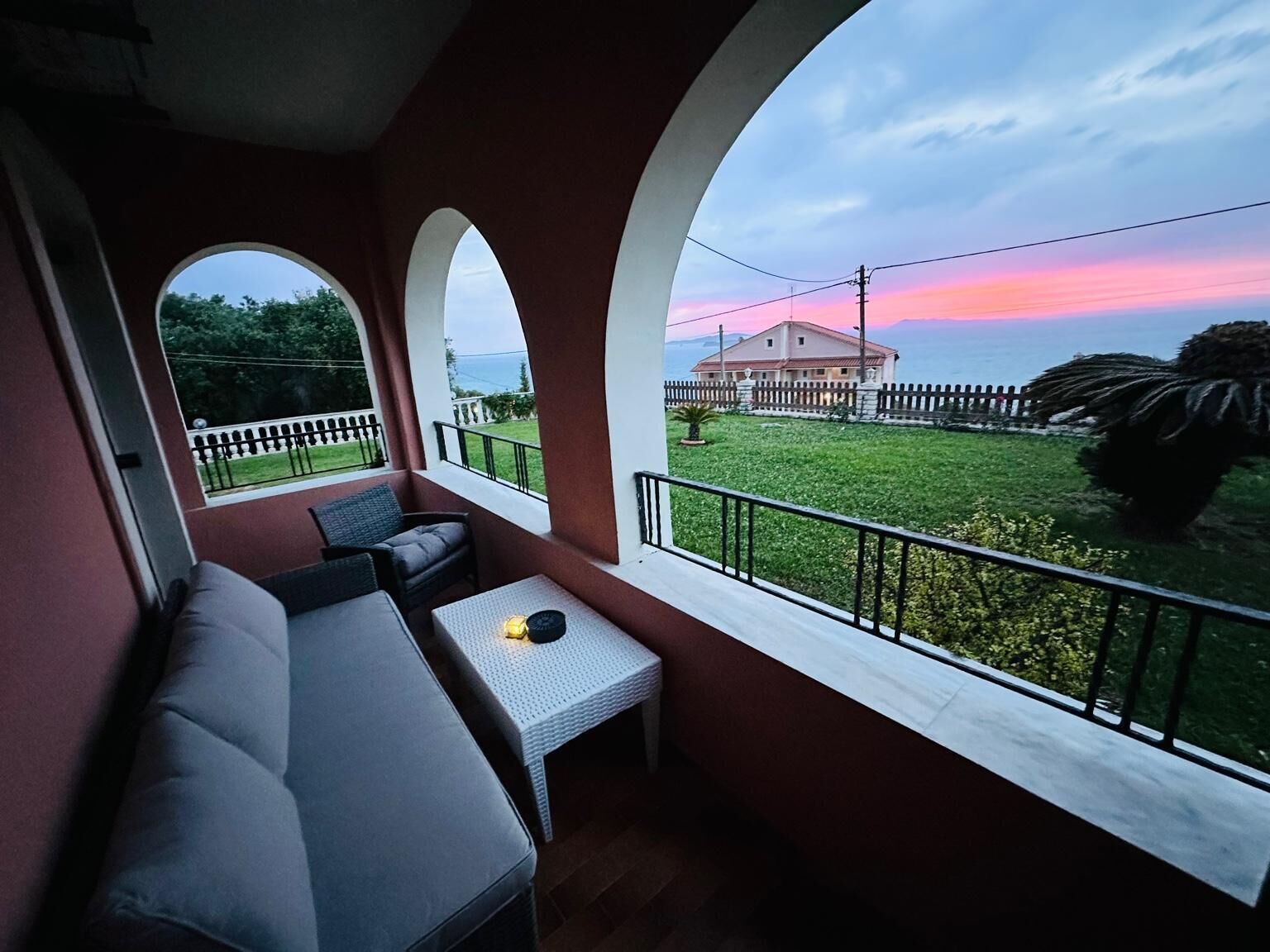 Ein Balkon mit einer Couch, einem kleinen Tisch und Blick auf ein Haus und Grünflächen.