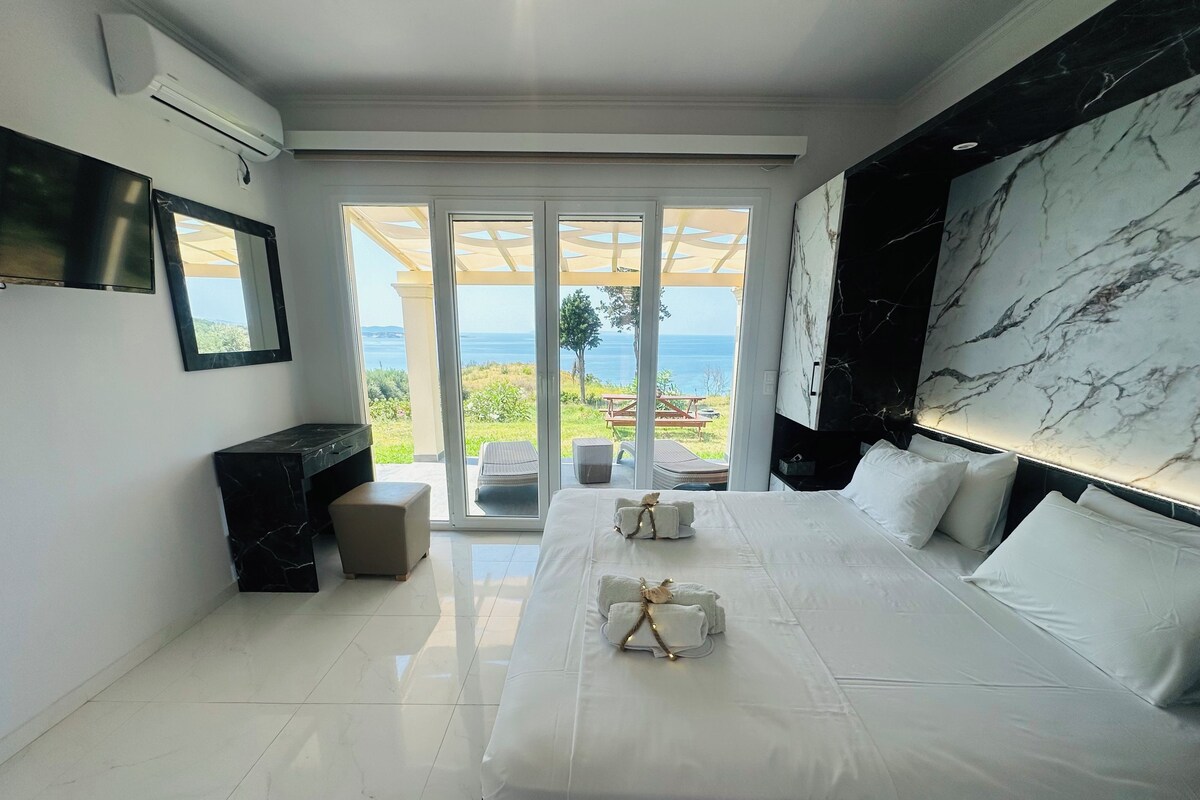 A modern hotel room with two beds, a marble headboard, a large window with a view of the sea, and a flat-screen TV.
