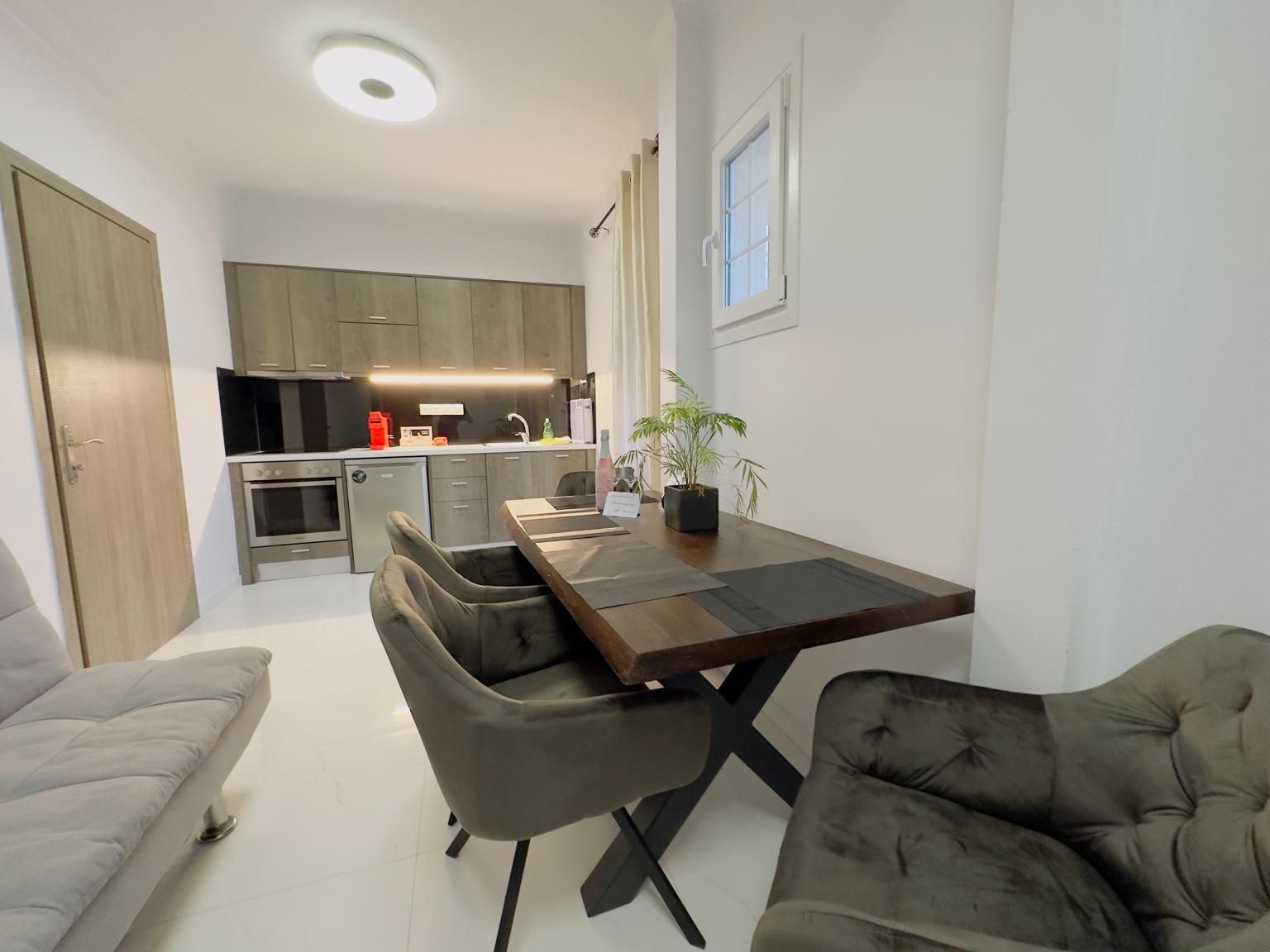 Une cuisine moderne avec des meubles de couleur sombre, un four encastré et un coin repas comprenant une table en bois et deux chaises grises recouvertes de tissu.