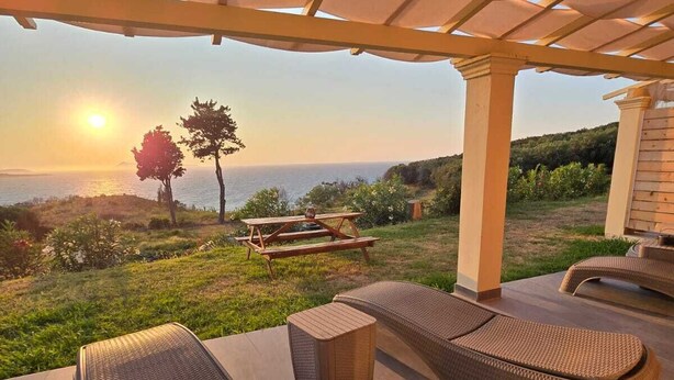 Un espacio de estar al aire libre cubierto con sillones de mimbre y una mesa de madera, con vistas panorámicas al océano y las colinas.