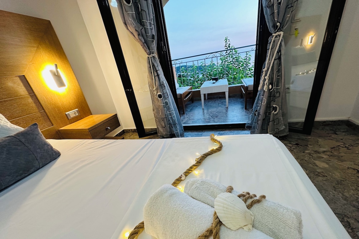 Habitación de hotel con cama, mesita de noche, lámpara de pared y vistas al exterior a través de cortinas abiertas.