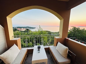 Un balcone con vista sul mare e sulle colline, arredato con un tavolo e due divani.