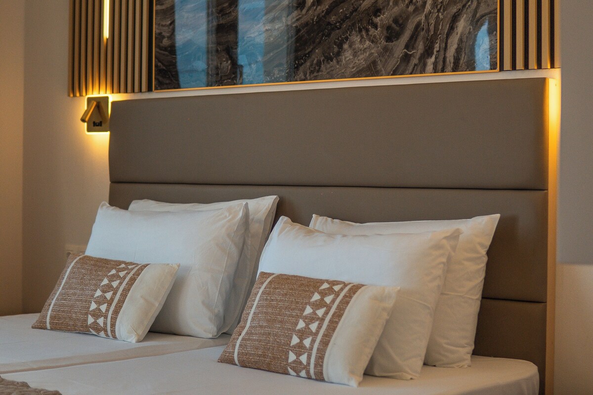 A neatly made bed with white linens and patterned pillows, a headboard with integrated lighting, and a large abstract painting above.