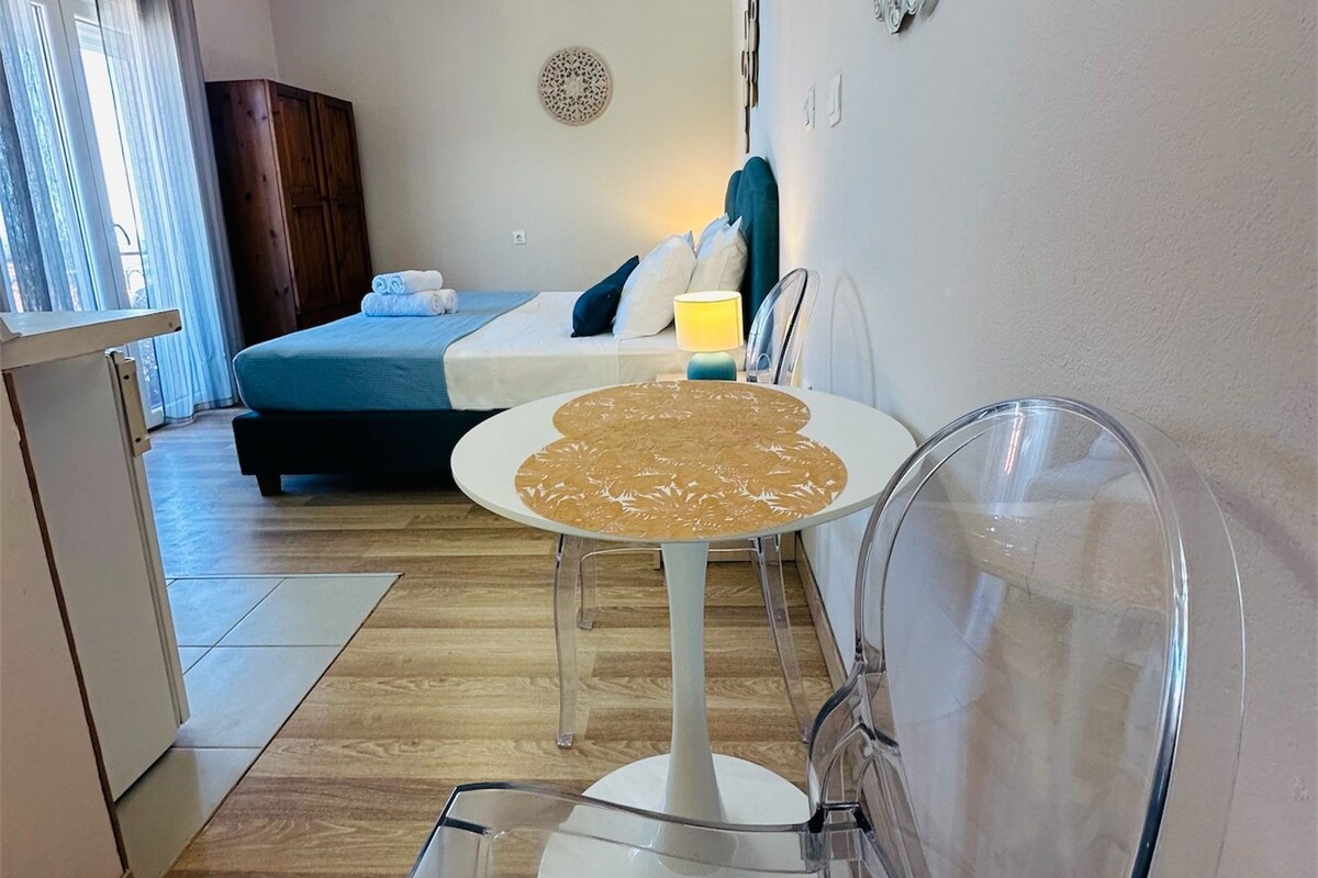 A hotel room with a bed, a dining table with chairs, a small kitchen area, and a decorative mirror on the wall.