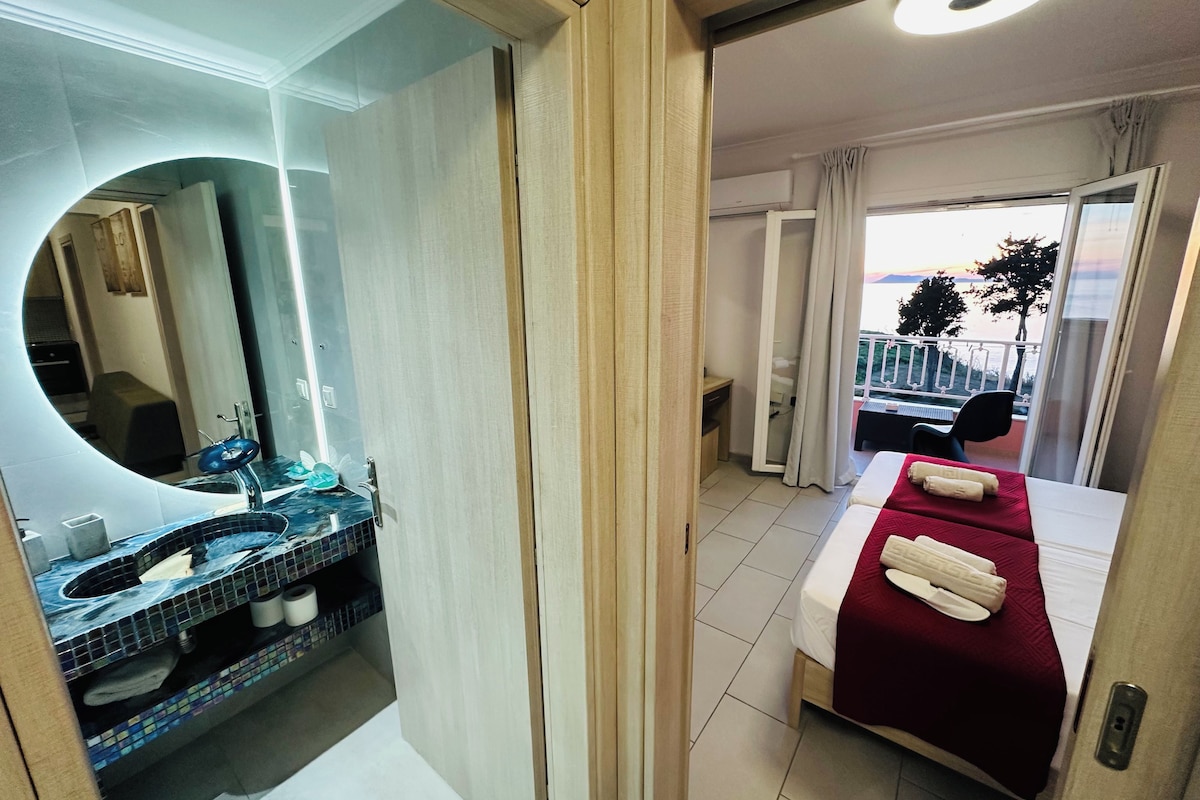 Habitación de hotel con cama, baño con lavamanos y espejo, y balcón con vistas.