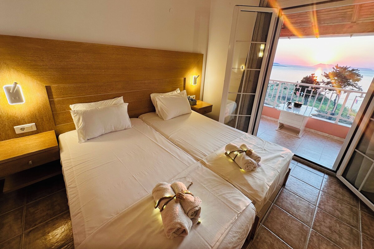 A hotel room with two beds, a wooden headboard, bedside lamps, and a balcony with a view of the sunset.