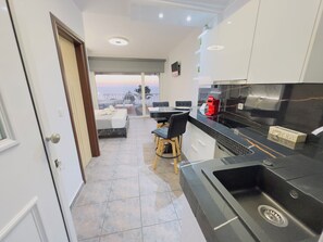 Una cocina moderna con encimera negra, horno empotrado y fregadero. Hay una zona de comedor con mesa y sillas, además de un balcón con vistas.