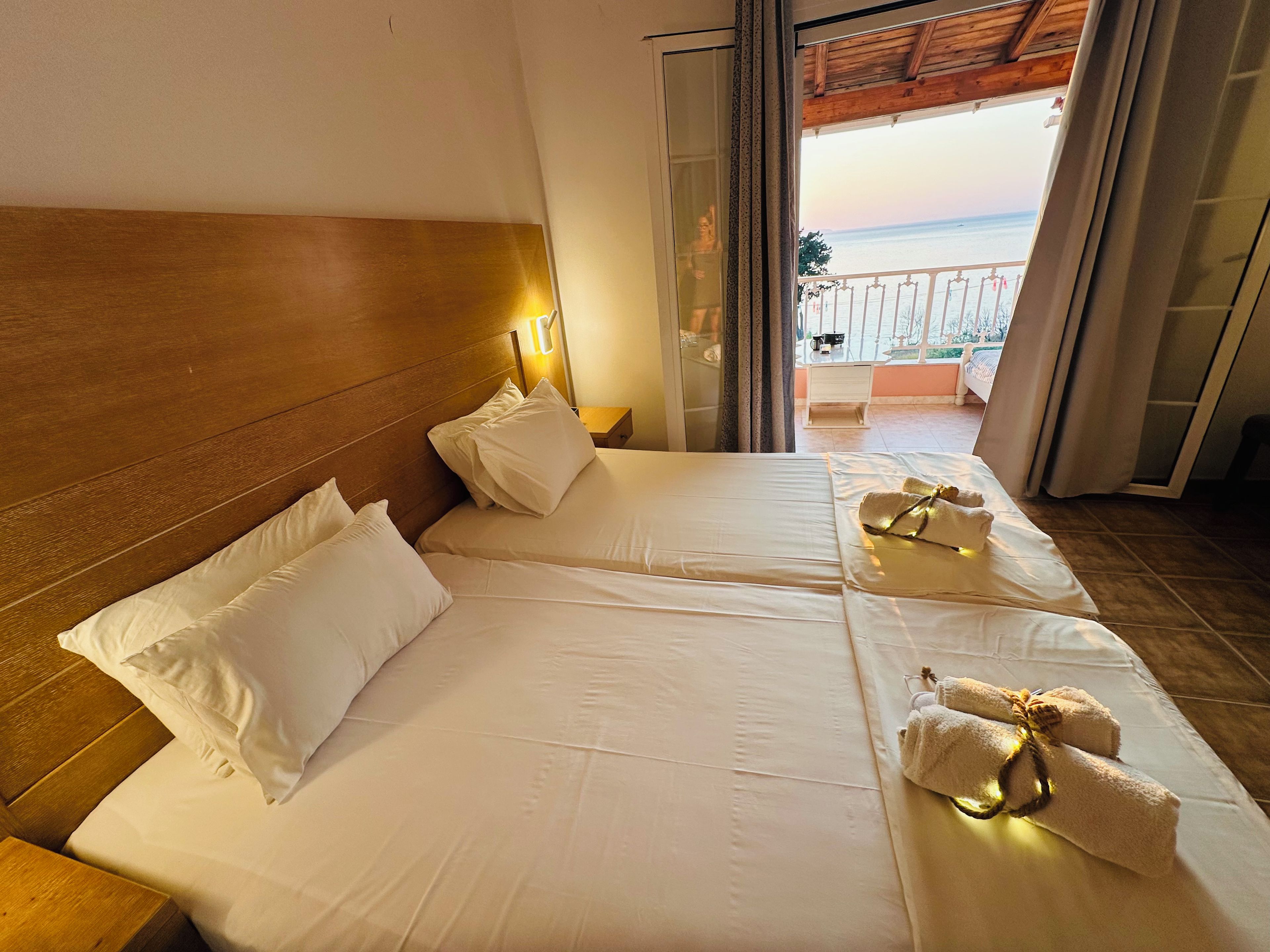 Habitación de hotel con dos camas, cabecero de madera, mesita de noche y vista al mar desde un balcón.