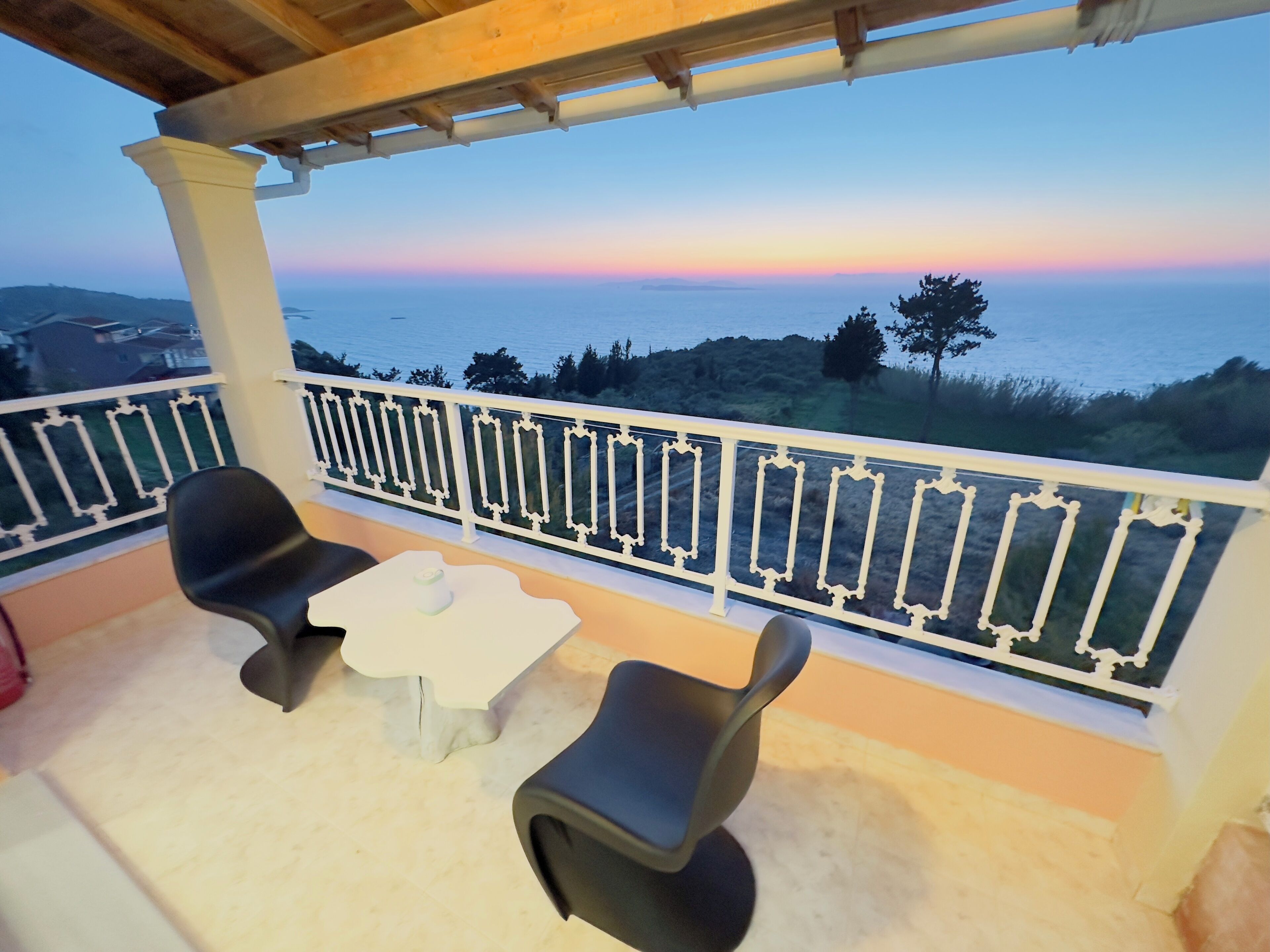 Ein Balkon mit zwei schwarzen Stühlen und einem kleinen Tisch, der einen malerischen Blick auf das Meer und die Hügel im Abendrot bietet.