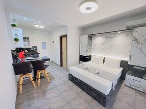 A modern hotel room with a large bed, a desk with two chairs, a small kitchen area, and a bathroom with a marble vanity.