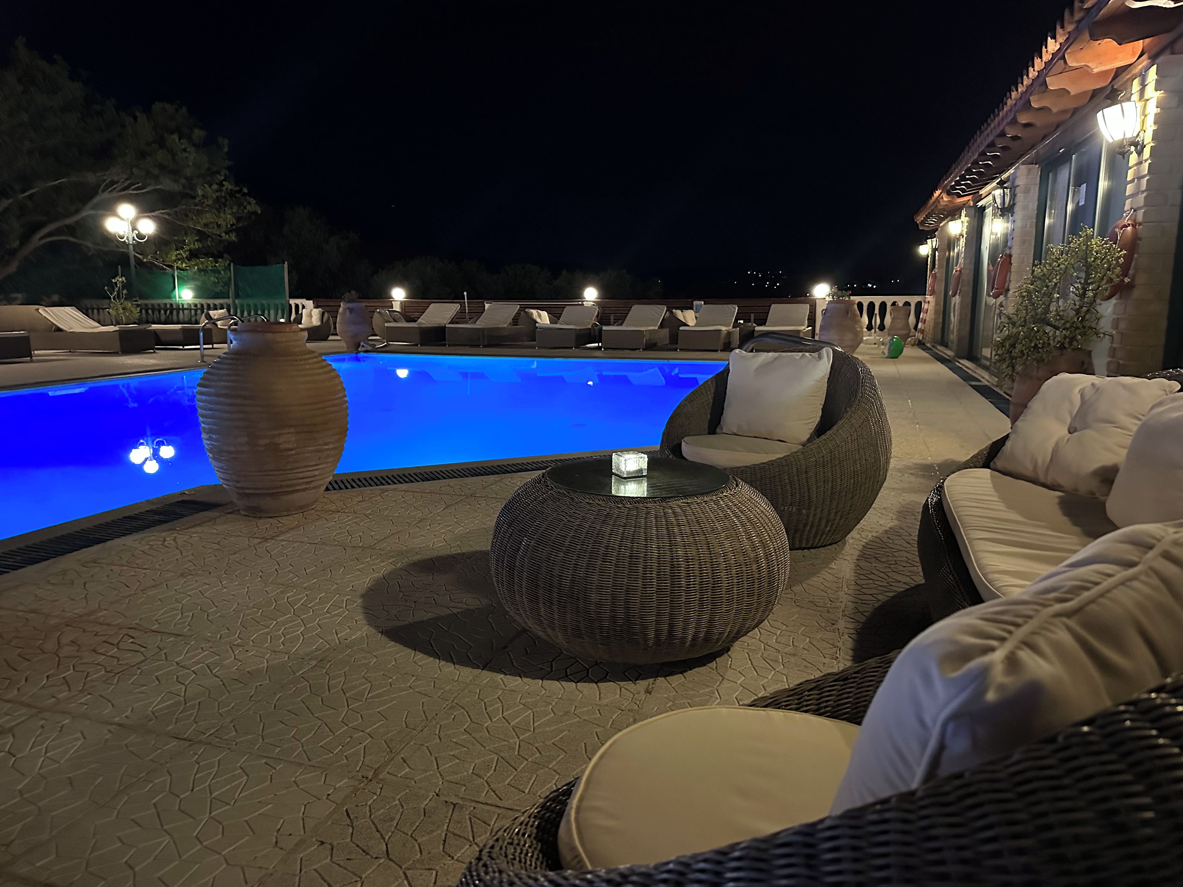 Een zwembadterras 's nachts met ligbedden, een ronde tafel en een grote vaas.