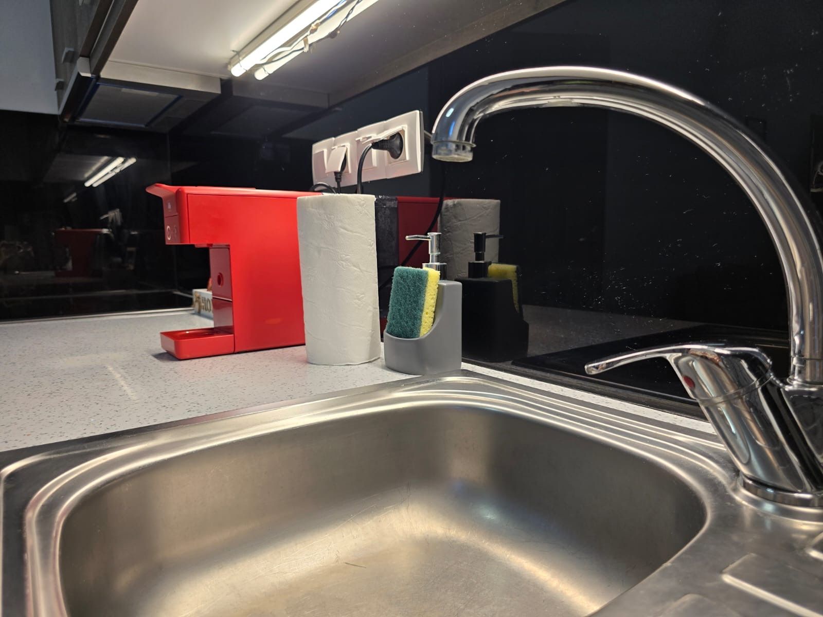 A modern kitchen with a stainless steel sink, a red countertop, and a mirror reflecting the space.