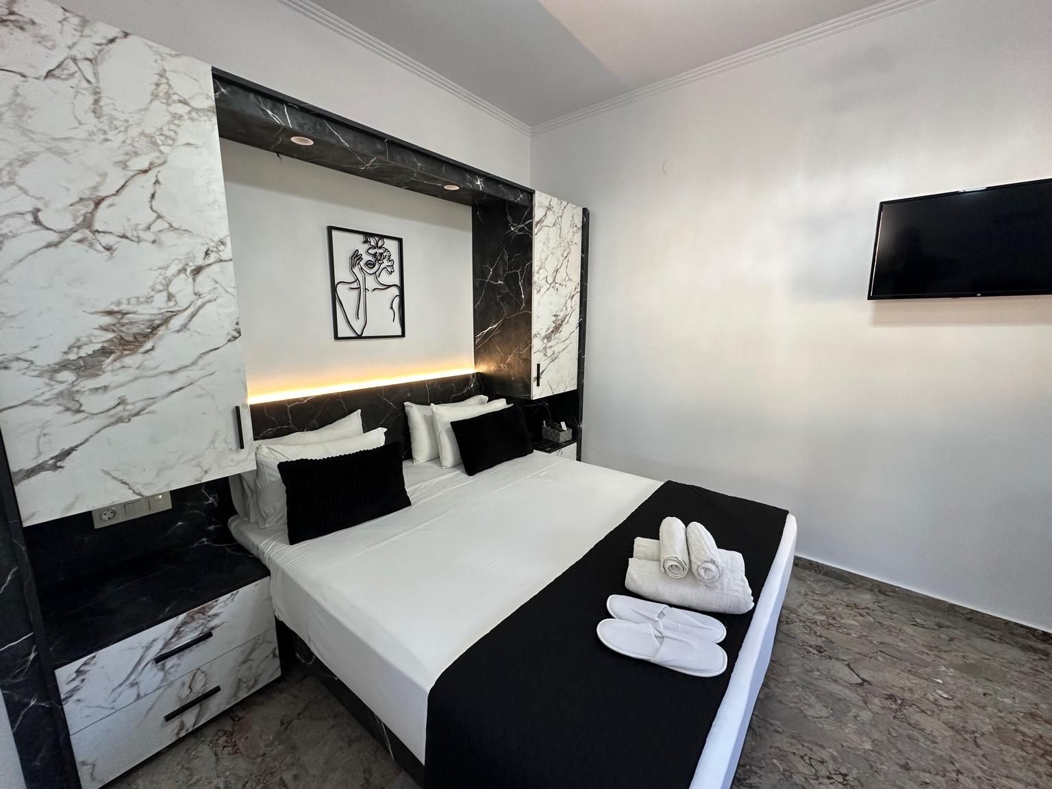 Une chambre d’hôtel moderne avec une tête de lit en marbre, un téléviseur à écran plat, un lit soigneusement fait avec du linge de lit blanc, et une paire de chaussons posée sur le lit.
