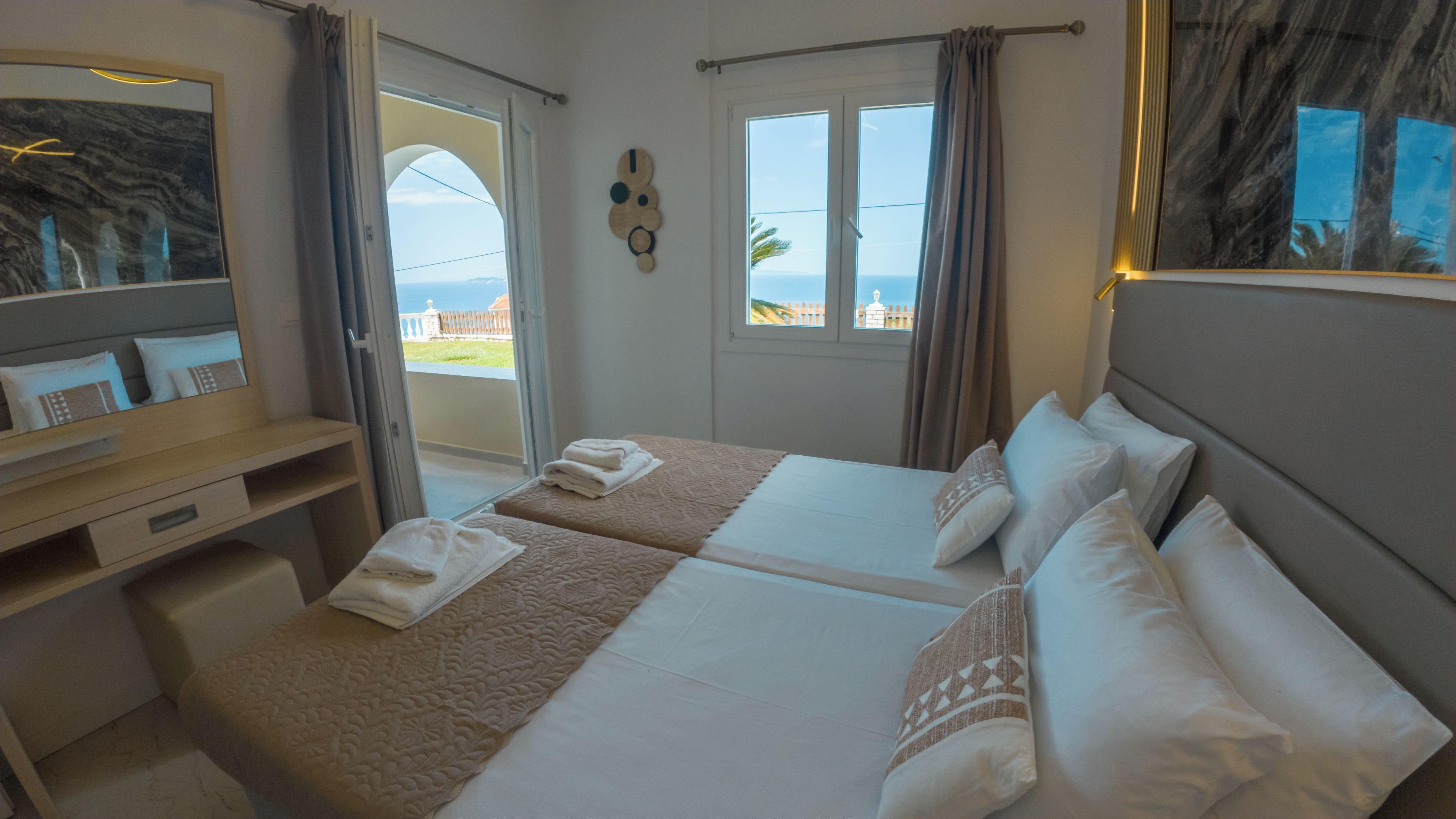 A hotel room with two beds, a desk, a mirror, and a view of the sea.