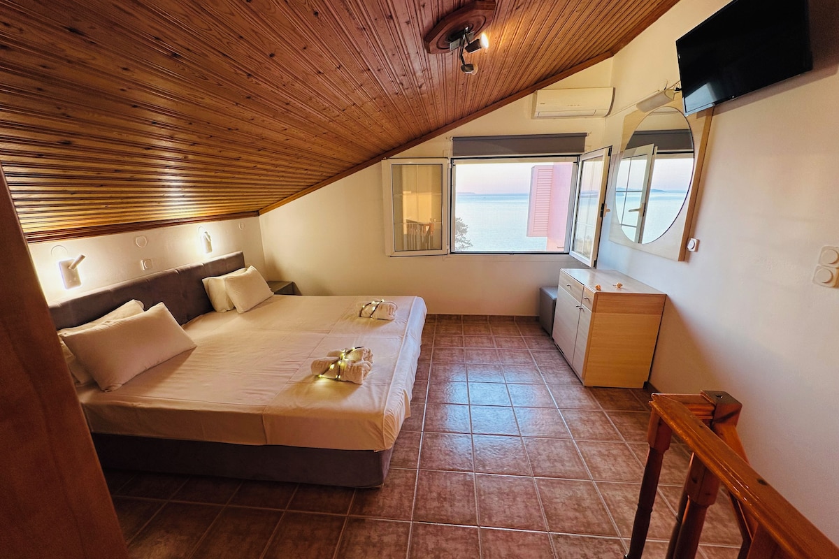 Un dormitorio con techo de madera inclinado, una cama con ropa blanca, una mesita de noche, un aparador y una ventana con vistas al mar.