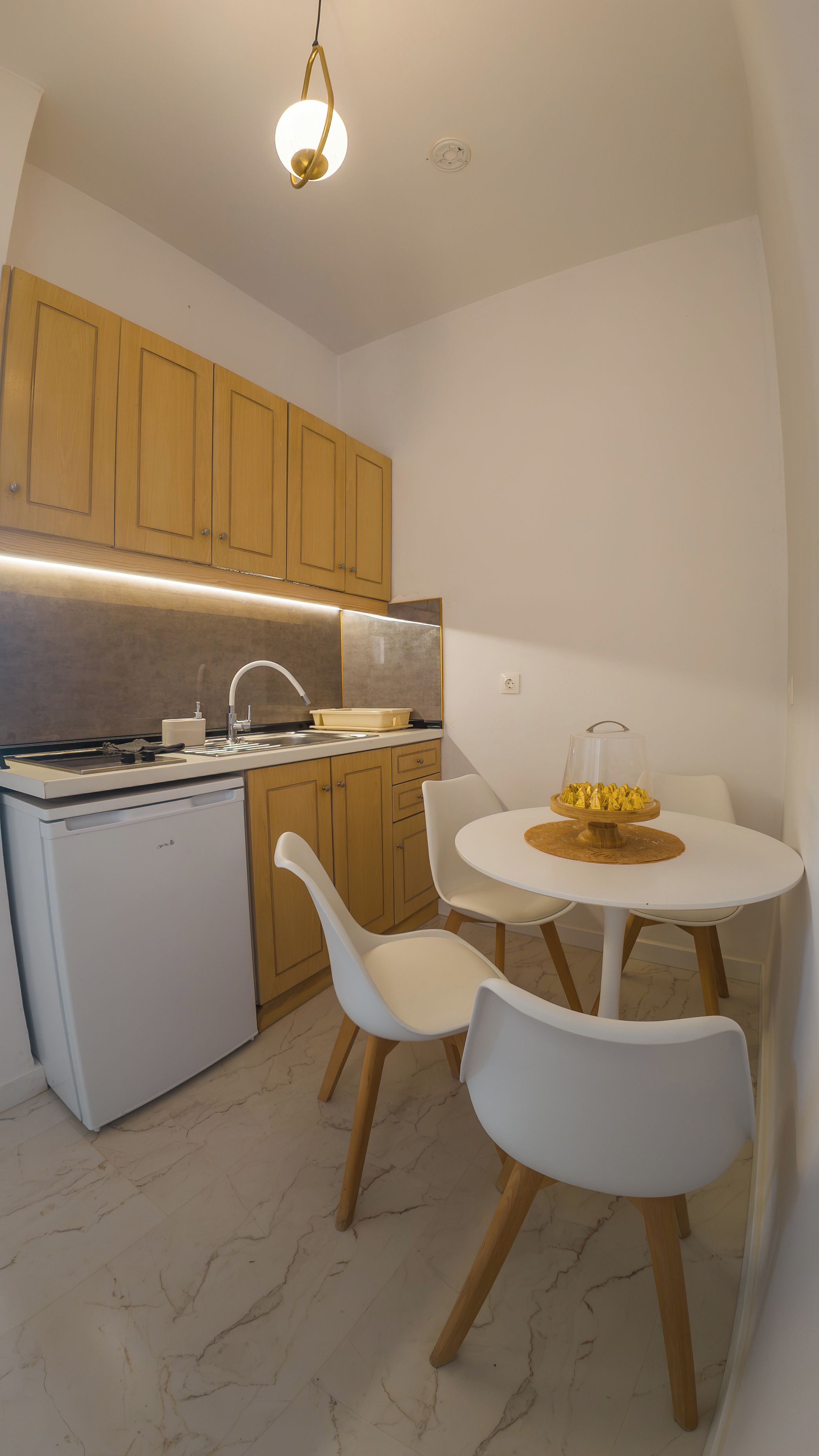Cocina compacta con armarios de madera, encimera blanca, lavavajillas integrado y una mesa redonda con dos sillas blancas.