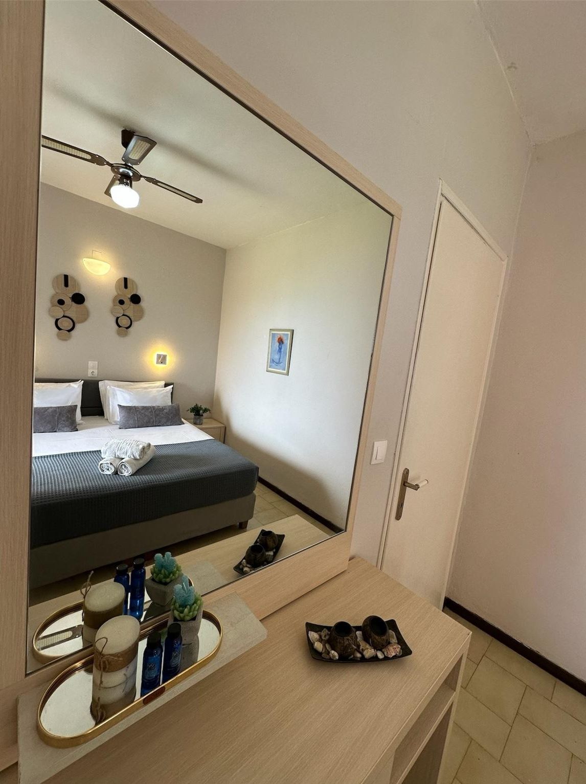 Hotelkamer met een grote spiegel, een bed met witte lakens, een plafondventilator en een klein nachtkastje met een lamp en decoratieve objecten.