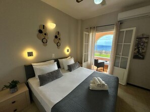 A hotel room with a bed, bedside table, wall decor, and a view of the sea through an open door. - Nikos Apartments Corfu (Corfu)