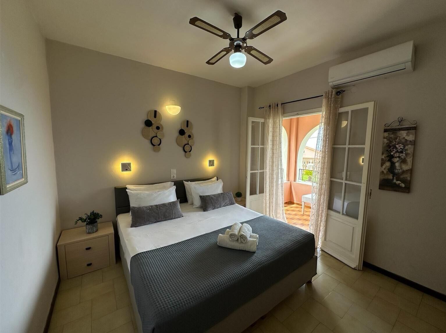 Ein Schlafzimmer mit Bett, Nachttischen, Deckenventilator, Wandleuchten, einer Tür zu einem Balkon und einem Wandbild.