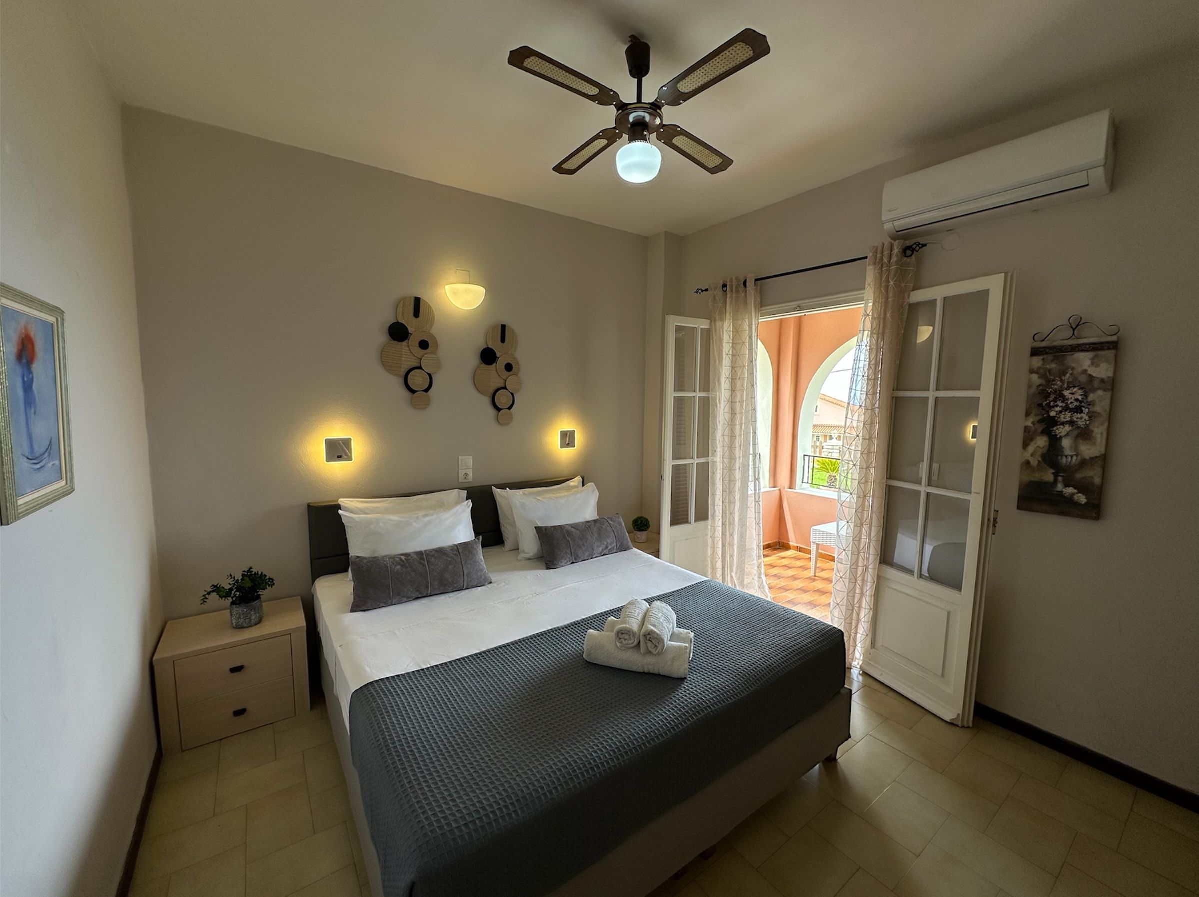 Une chambre à coucher comprenant un lit, des tables de chevet, un ventilateur de plafond, des appliques murales, une porte donnant sur un balcon et un tableau accroché au mur.
