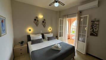 Une chambre à coucher comprenant un lit, des tables de chevet, un ventilateur de plafond, des appliques murales, une porte donnant sur un balcon et un tableau accroché au mur.