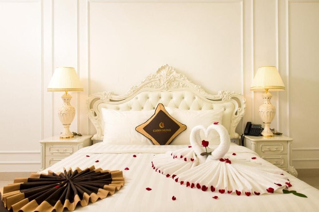 Ein luxuriöses Bett mit weißem Kopfteil, einem Zierkissen und einer schwanenförmigen Anordnung aus weißen und roten Rosenblüten.