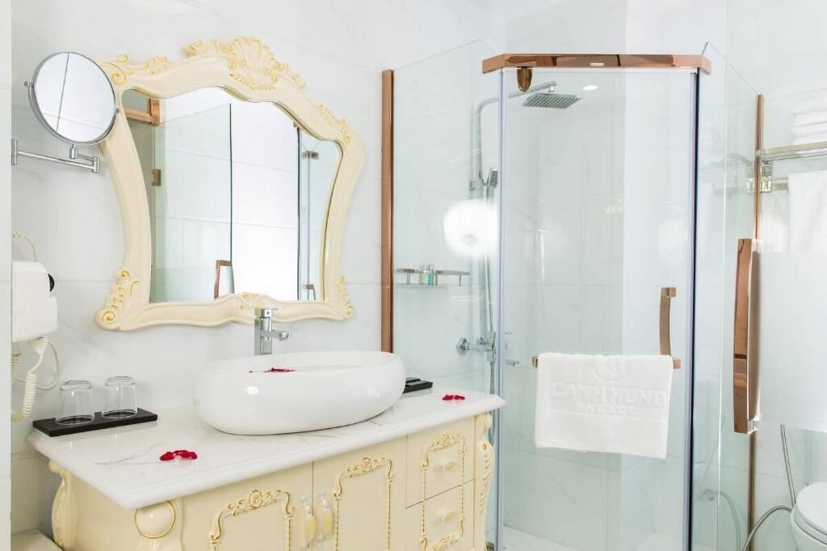 Un baño con lavamanos blanco, un espejo grande con detalles dorados ornamentales y una cabina de ducha de vidrio.