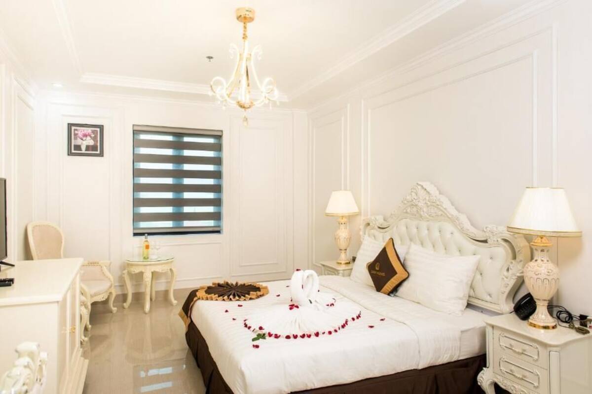 Habitación de hotel con cama, mesitas de noche, escritorio, silla, candelabro y ventana con persianas.