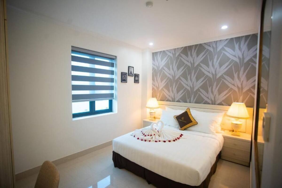 Habitación de hotel con una cama bien hecha, dos lámparas de noche, una ventana con persianas y una pared con un diseño de hojas.