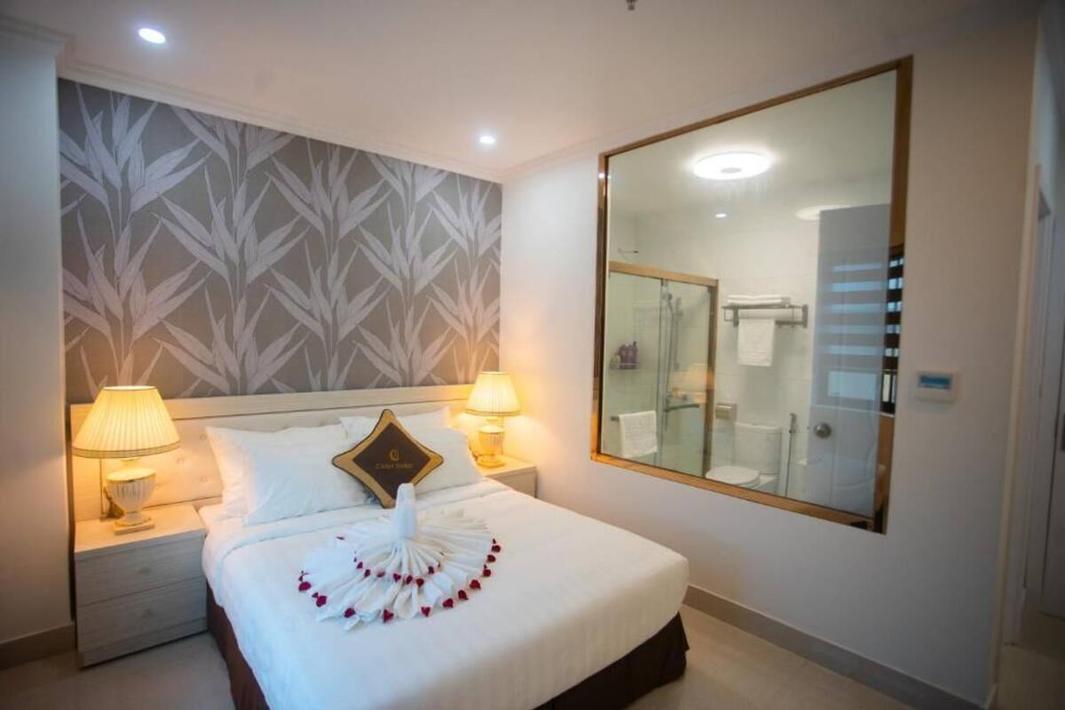 Habitación de hotel con una cama bien hecha, dos lámparas de noche, un espejo grande y un baño visible a través de la puerta de vidrio.