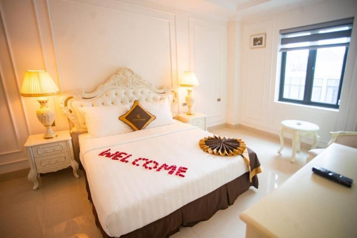 Una habitación de hotel con una cama grande, dos lámparas de noche, una mesita redonda y un ventanal con persianas.