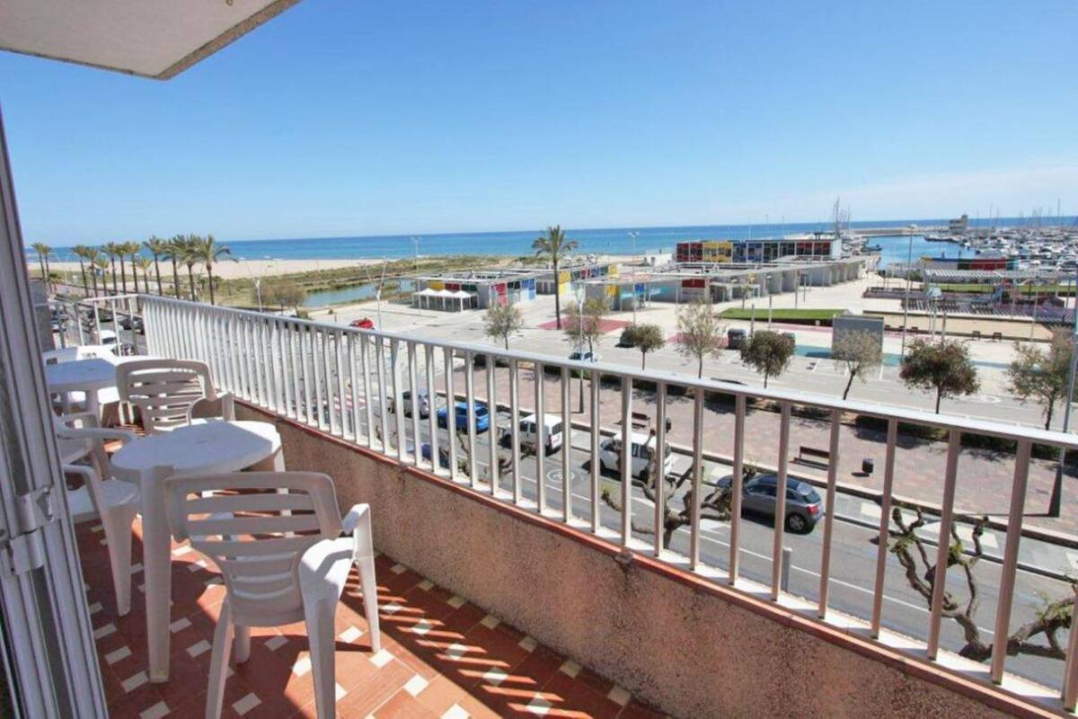 A balcony with white plastic chairs and a table, overlooking a marina with boats and a beach.