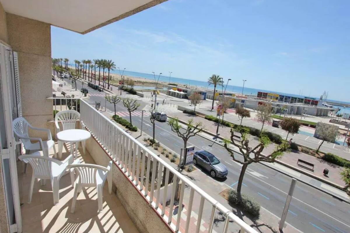 A balcony with white plastic chairs and a table, overlooking a street with palm trees and a beach in the background.