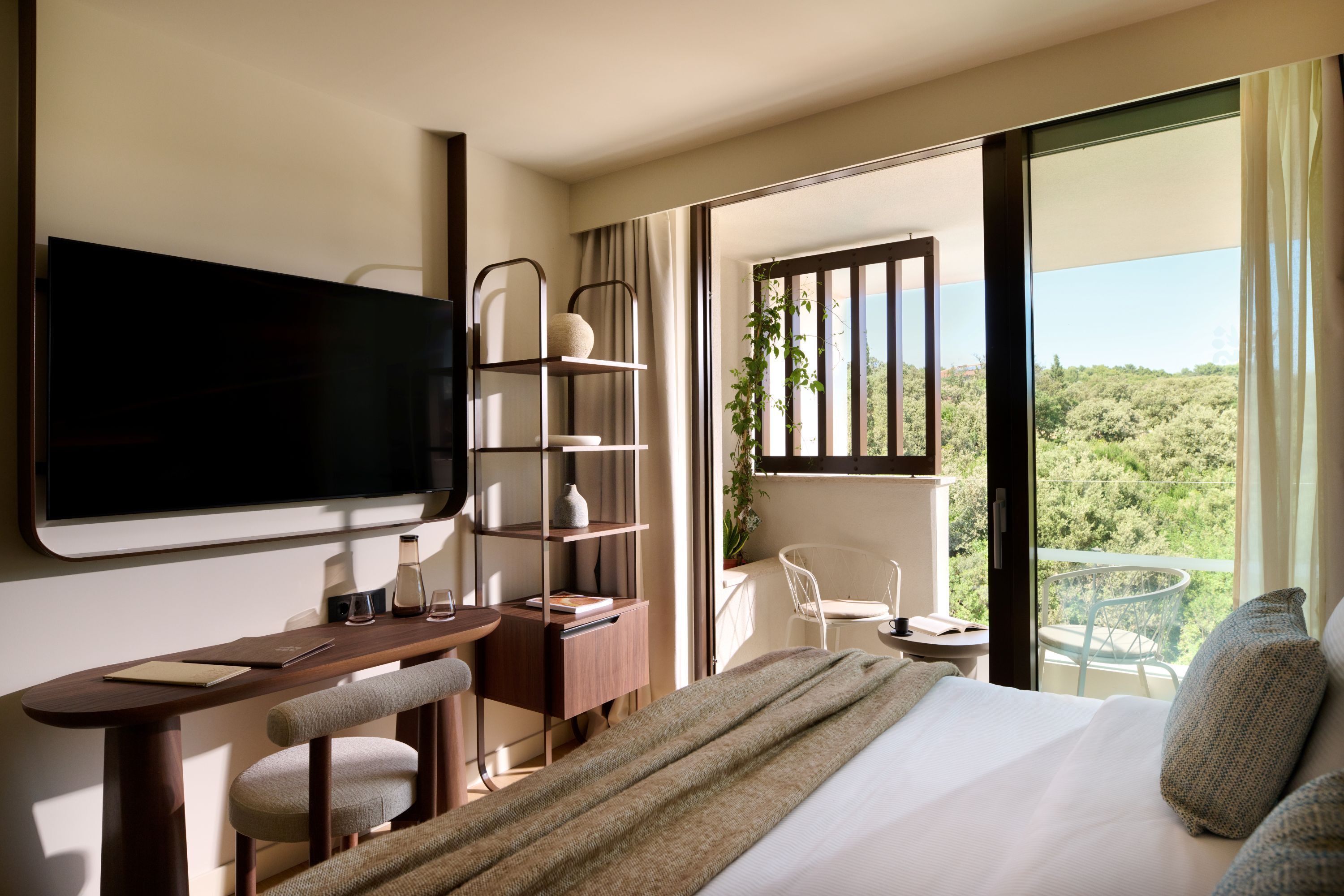 A modern hotel room with a large bed, a desk with a chair, a TV, and a balcony with a view of the greenery.
