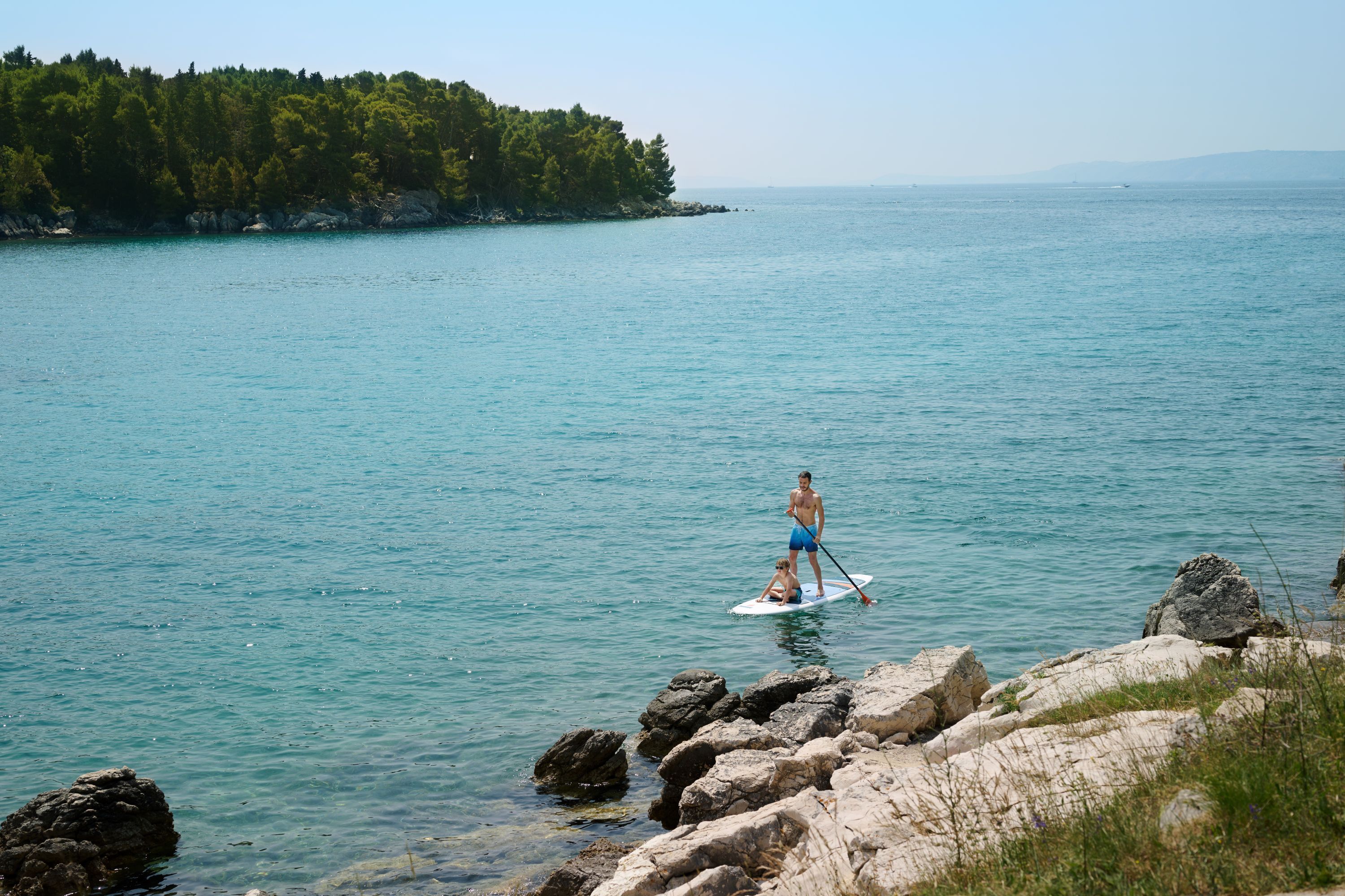 Una persona fa paddleboarding su un'acqua calma con una riva rocciosa e un'isola boscosa sullo sfondo.