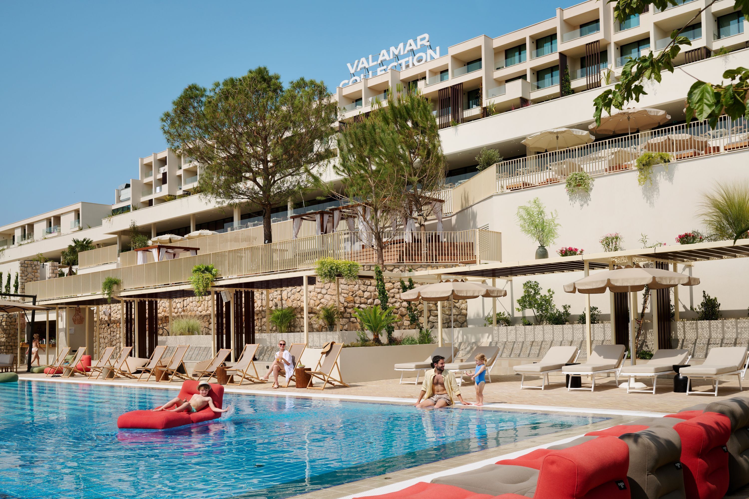 Ein Hotelpoolbereich mit Liegestühlen und Sonnenschirmen, ein klarer Himmel und ein Gebäude mit Balkonen.