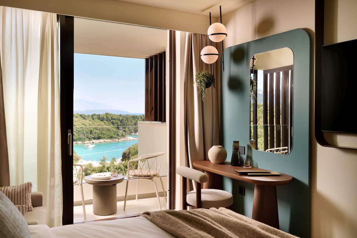 A modern hotel room with a balcony, a view of a coastal area, a bed, a desk, and a chair.