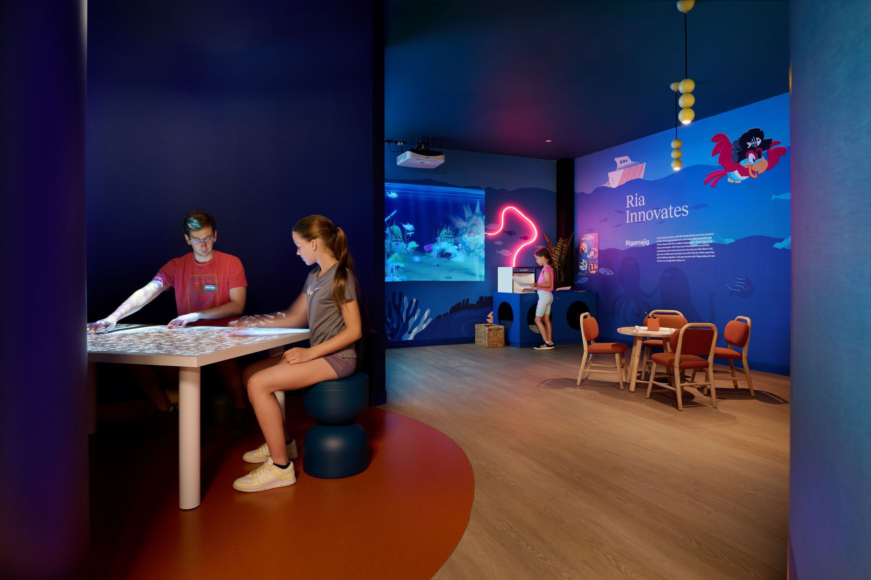 Een moderne kamer met een groot interactief scherm, een tafel met een laptop, en een zithoek met stoelen en een kleine tafel.