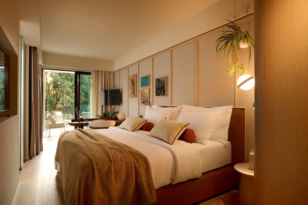 A modern hotel room with a large bed, a TV, a desk, and a view of the outdoors.