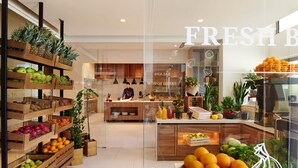 A store interior with wooden shelves stocked with fruits and vegetables, a central counter with fresh produce, and a visible sign reading "FRESH B".