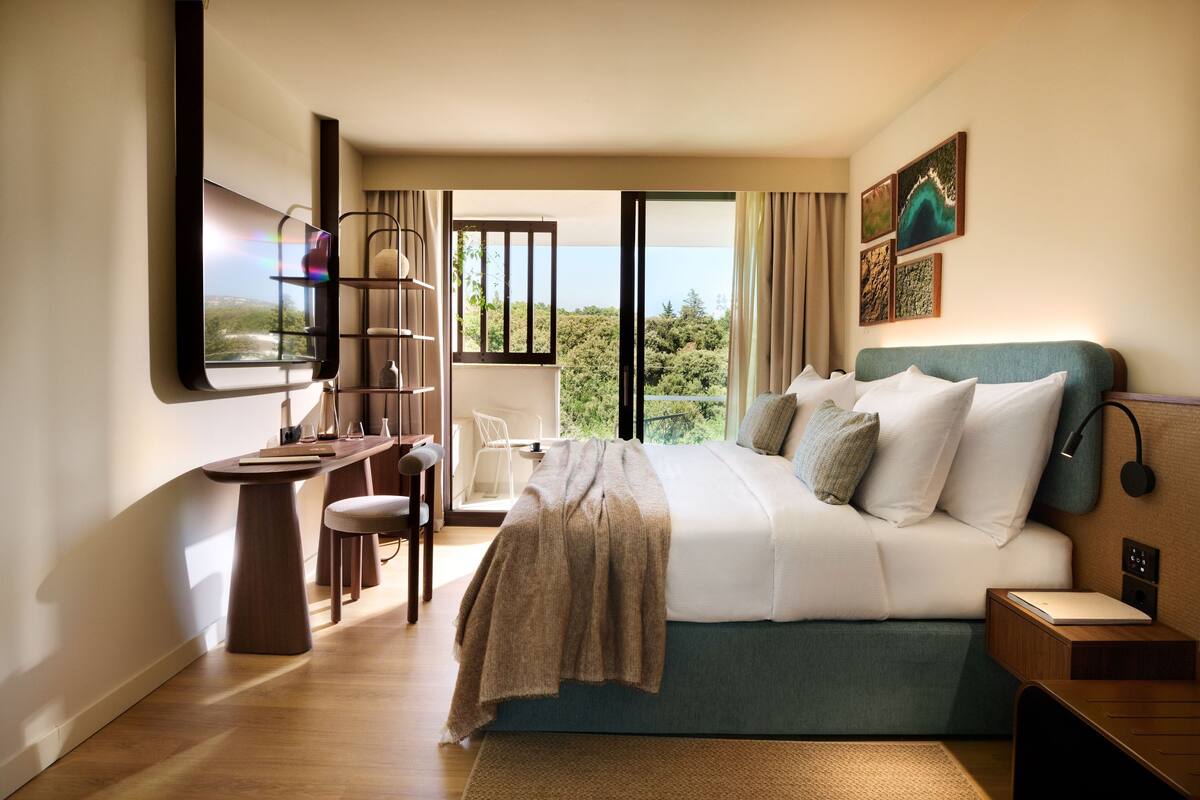 A modern hotel room with a bed, a desk, and a chair. There is a large window with a view of greenery.