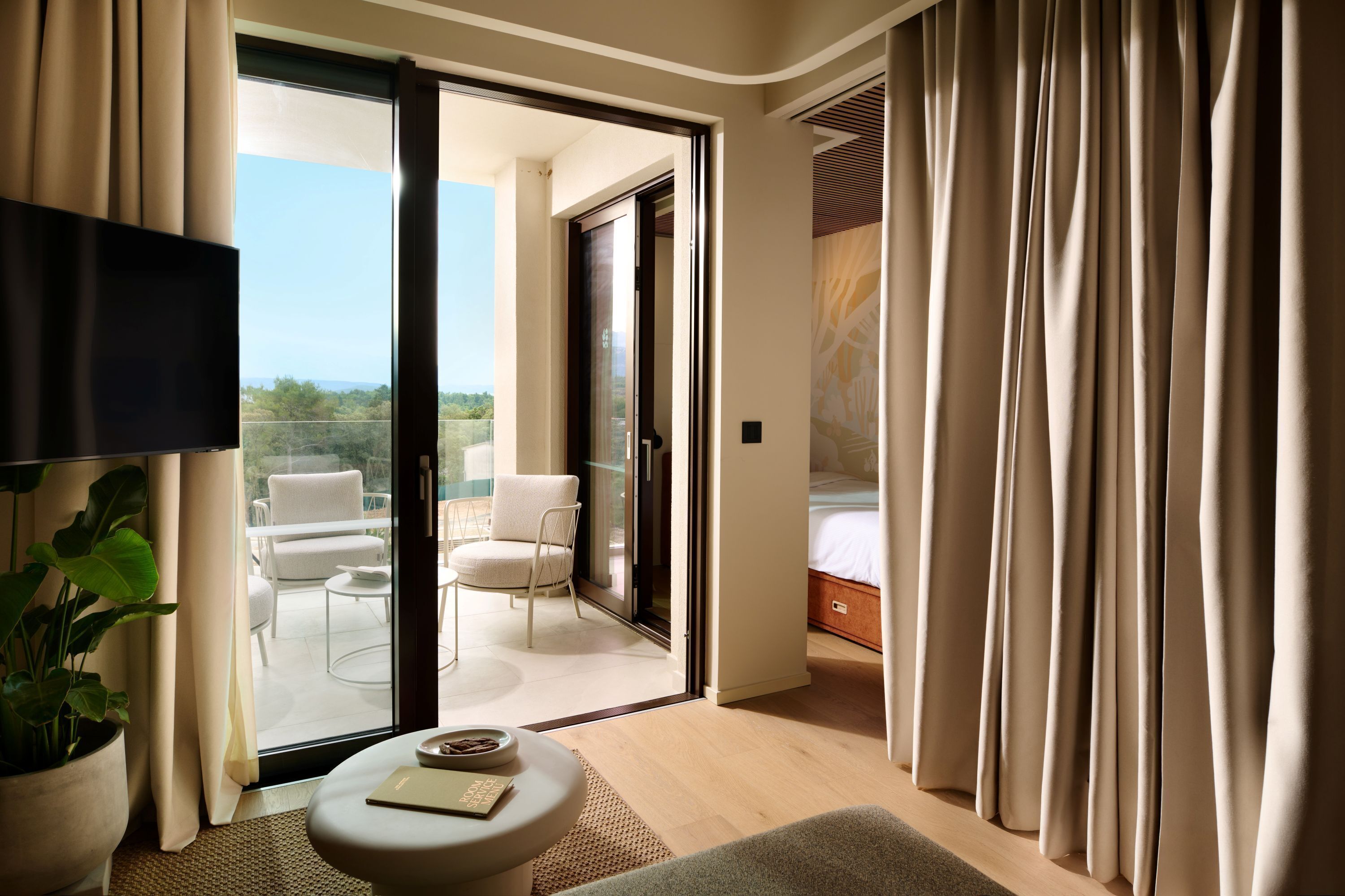 A hotel room with a balcony, a flat-screen TV, a potted plant, a round table, and a bed.