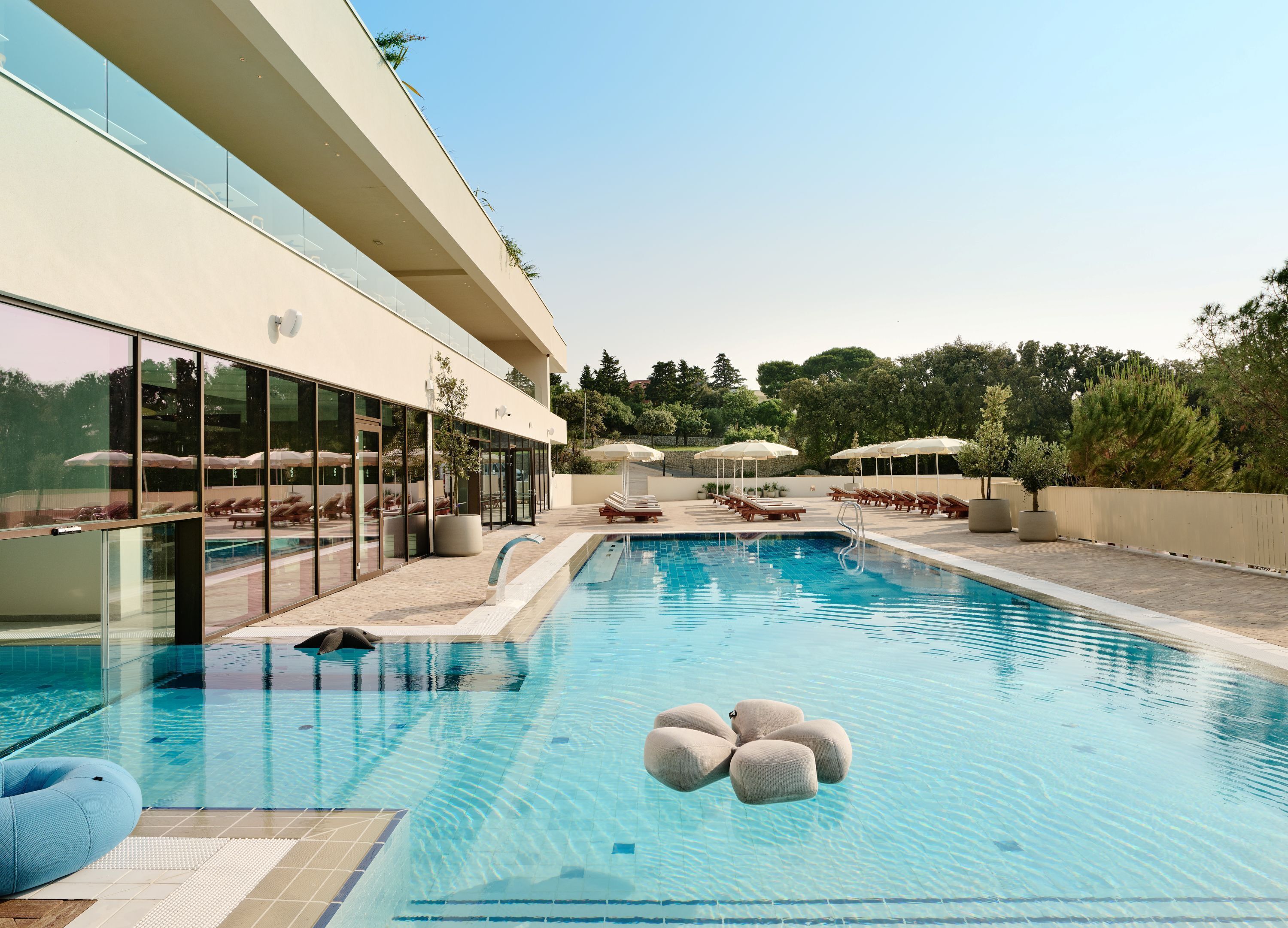 Zona de piscina moderna de hotel con paredes de vidrio transparente, sillas de descanso y sombrillas.