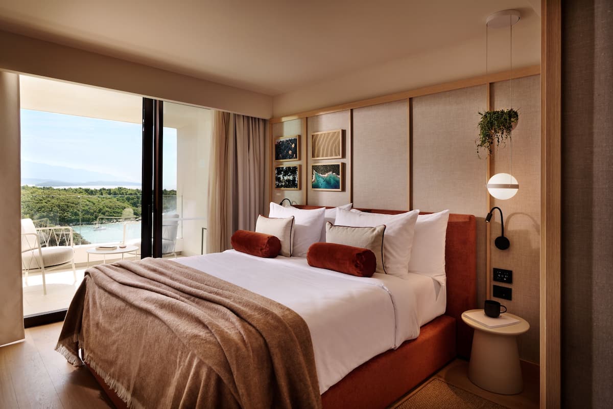 A hotel room with a bed, a bedside table, a view of the outdoors, and a wall with artwork.