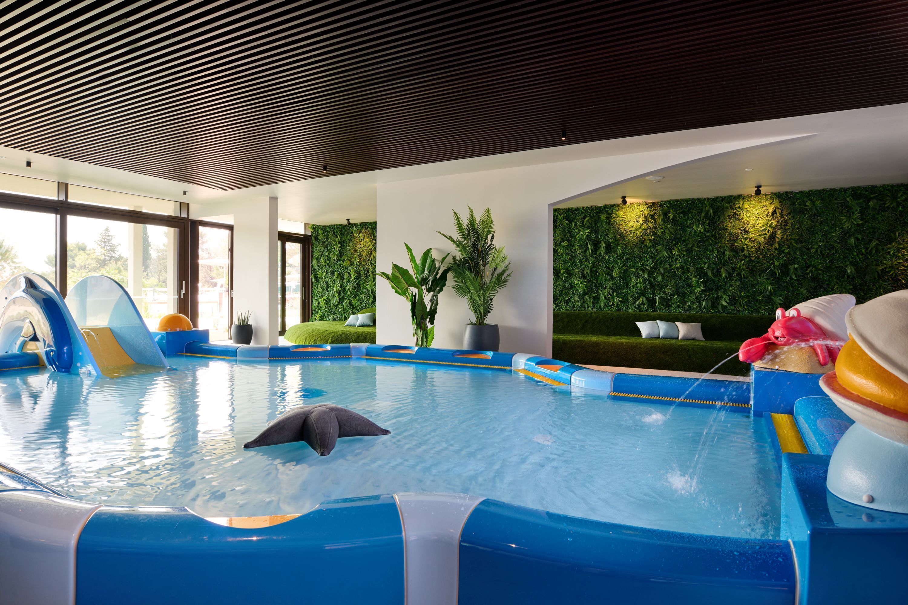 Una piscina con scivoli e un delfino, circondata da pareti verdi e ampie finestre.