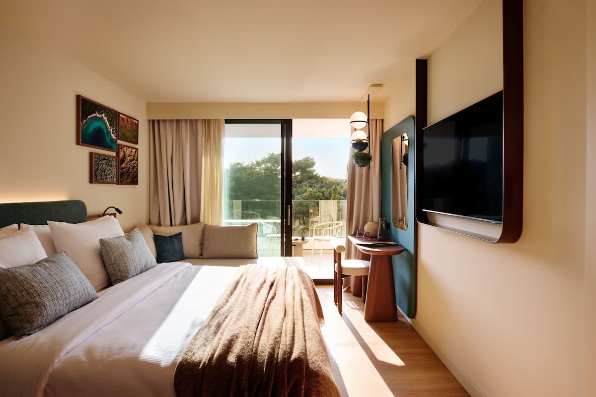 A hotel room with a large bed, a flat-screen TV, a desk with a chair, and a balcony with a view of trees.