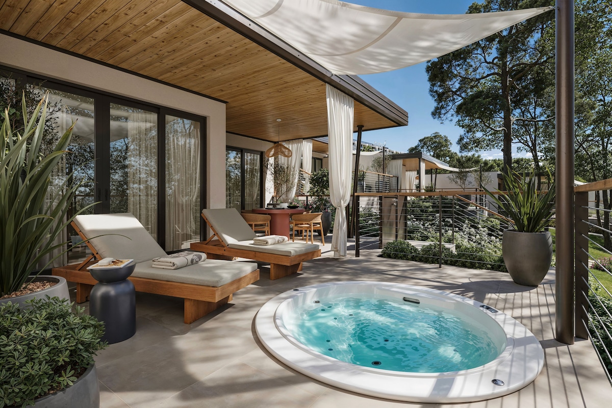 A modern outdoor hot tub area with lounge chairs, a dining table, and a view of the surrounding landscape.