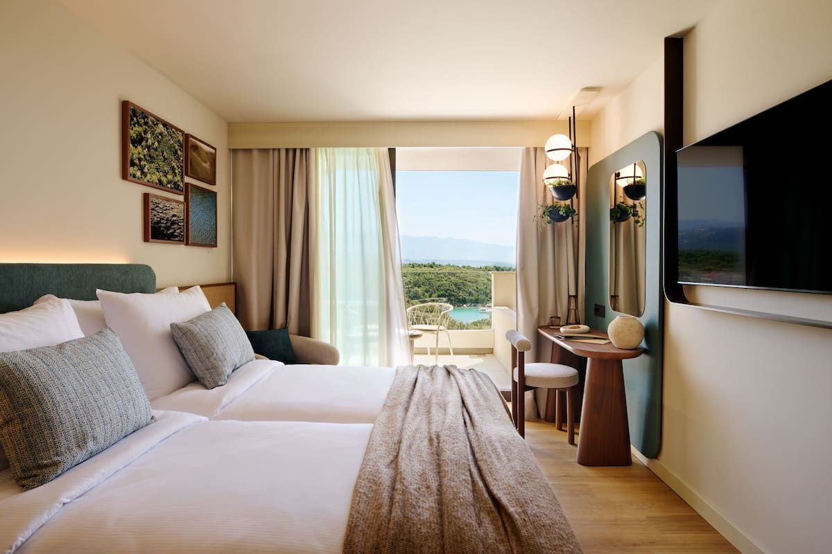 A hotel room with two beds, a balcony with a view, a TV, and a small table with chairs.