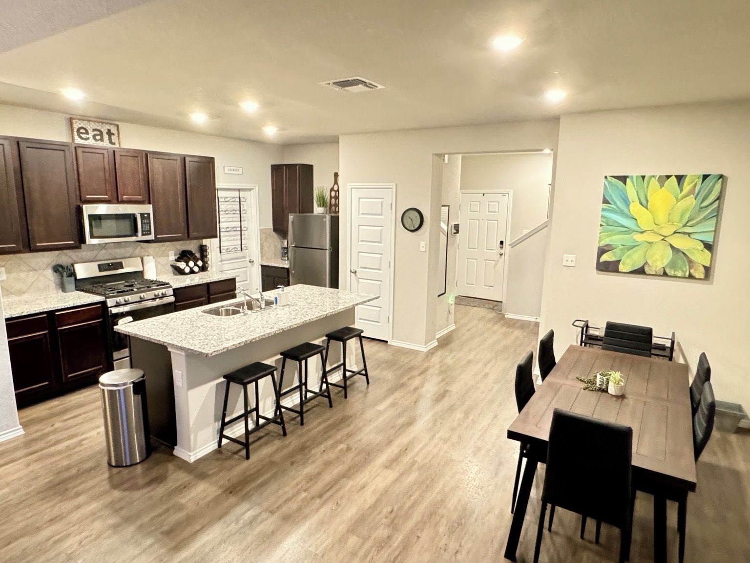 A modern kitchen with a central island, dark wooden cabinets, stainless steel appliances, and a dining area with a wooden table and chairs.