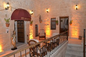 Interior de paredes de piedra con un toldo rojo, plantas en macetas y una estatua.